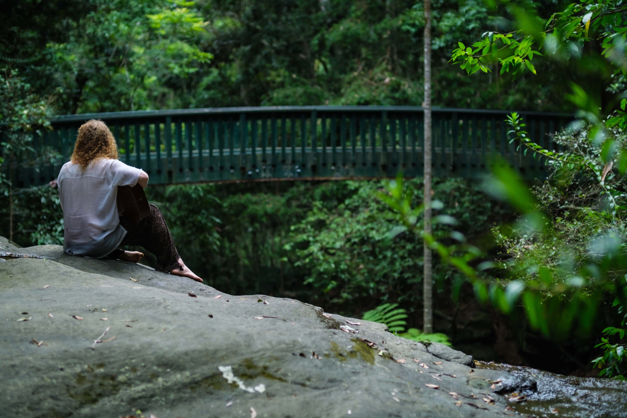 Alexander seated near a forest bridge, with his guitar.